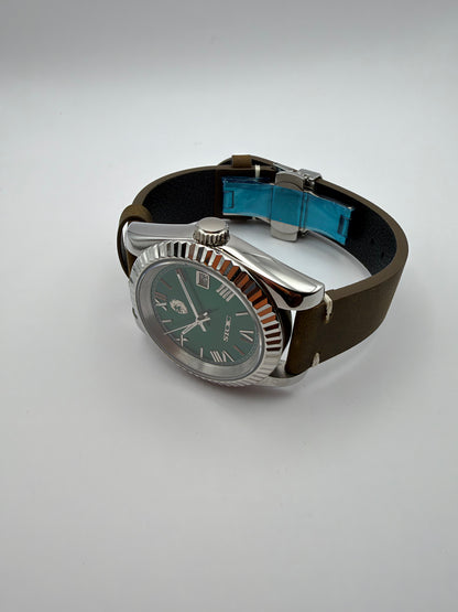 Verdant resting at an angle, showing its silver-tone case, brown strap and blue film on the folding clasp.